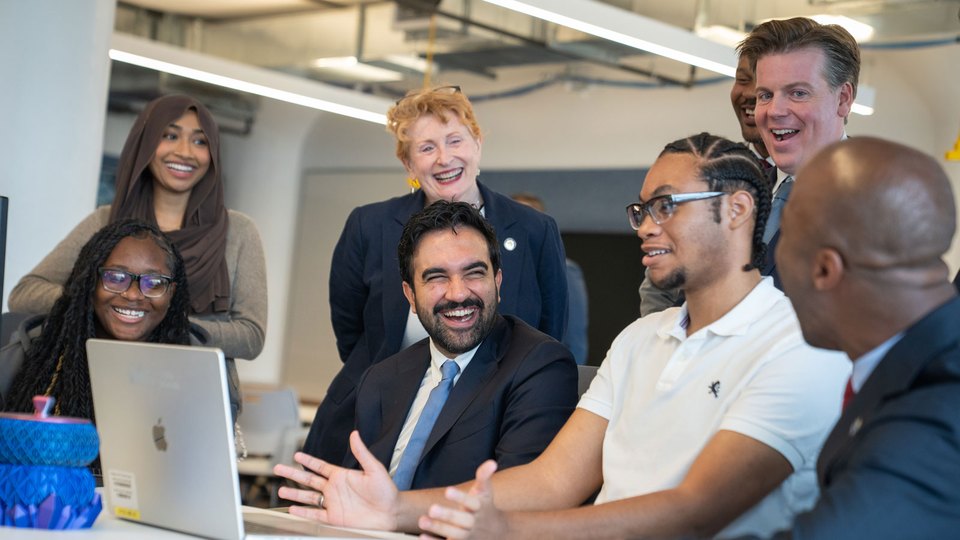 Mayor Mamdani seated with students and staff around an open laptop, mid-conversation.